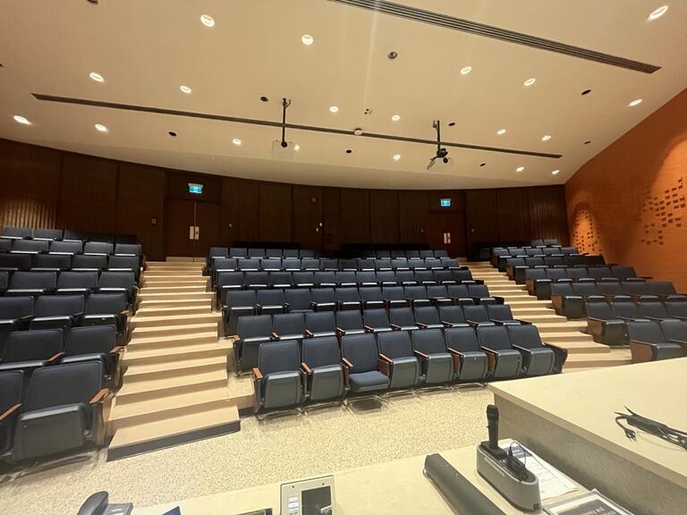 View of the classroom from the instructor’s podium. There are multiple rows of chairs for students, with tiered steps to access tables in the middle and back area of the classroom. This classroom is not accessible for instructors.