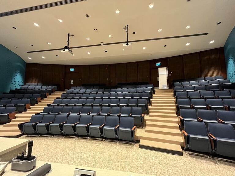View of the classroom from the instructor’s podium. There are six rows of chairs for students, with tiered steps to access tables in the middle and back area of the classroom. This classroom is not accessible for instructors.
