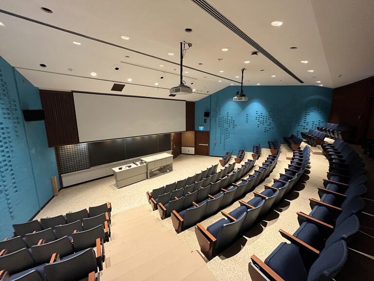 View of the classroom from a student seat near the rear of the room. In front are multiple empty rows of chairs. The instructor podium is at the front of the room, in the middle. The classroom has a long blackboard in the middle of the front wall. Two digital projectors on the ceiling, project onto a screen at the front of the room. This classroom is not accessible for students.