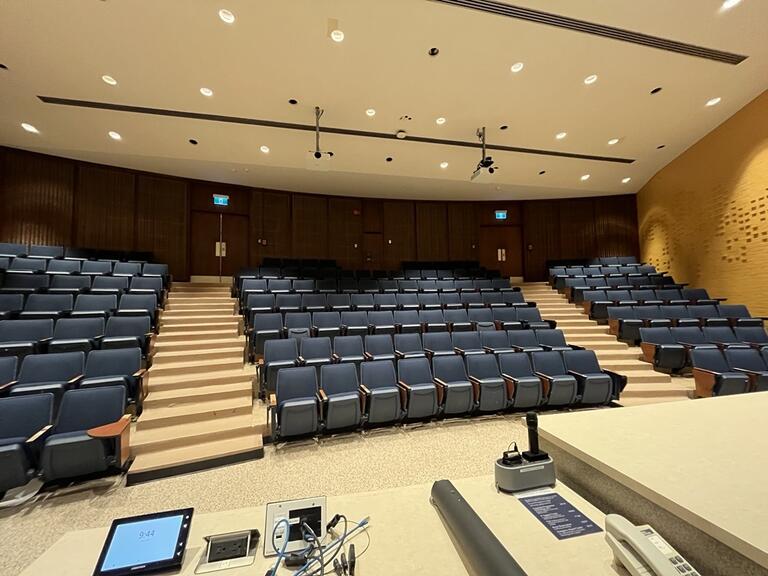 View of the classroom from the instructor’s podium. There are six rows of chairs for students, with tiered steps to access tables in the middle and back area of the classroom. The entryway door is located at the back of the room, on the right. This classroom is not accessible for instructors.