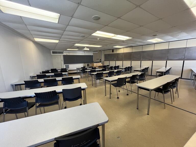 View of the classroom from a student seat near the rear of the room. In front are multiple rows of empty tables and chairs. The instructor podium is located at the front of the room on the right. The front of the classroom has a long blackboard in the middle of the front wall. A digital projector on the ceiling, projects onto a screen that extends from the ceiling. The entryway door is located at the front of the room, on the left. This classroom is accessible for students.