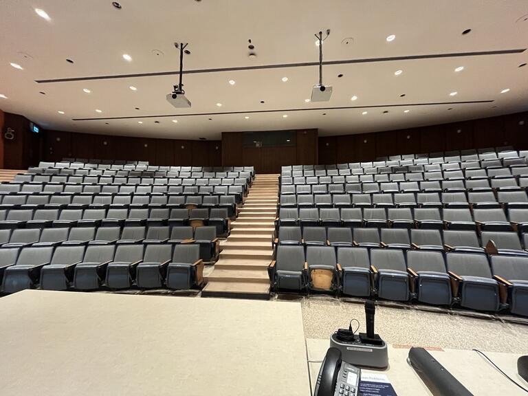View of the classroom from the instructor’s podium. There are multiple rows of chairs for students, with tiered steps to access tables in the middle and back area of the classroom. This classroom is not accessible for instructors.