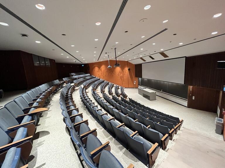 View of the classroom from a student seat near the rear of the room. In front are multiple empty chairs. The instructor podium is at the front of the room in the middle. The front of the classroom has a long blackboard in the middle of the front wall. Two digital projectors on the ceiling, project onto a screen that extends from the ceiling. The entryway door is located at the front of the room, on the right. This classroom is not accessible for students.