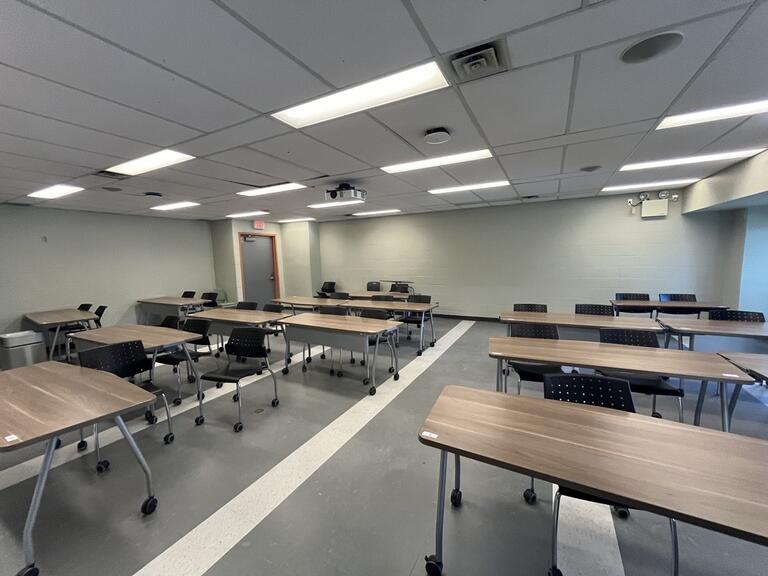 View of the classroom from the instructor's podium. In front are rows of movable tables and chairs. The door is located at the left. Chairs and tables are movable to fit the instructors needs. The classroom is accessible for instructors.