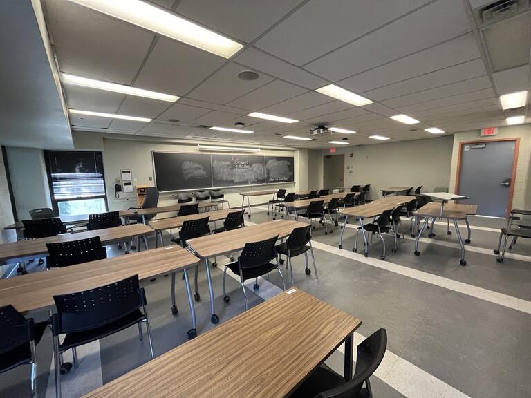 : View of the classroom from a student seat near the rear of the room. In front are rows of movable tables and chairs. The instructor’s wall podium is located at the front of the room on the left. A blackboard is located on the front wall. A digital projector on the ceiling, projects onto a screen that extends from the ceiling. This classroom is accessible for students and instructors.