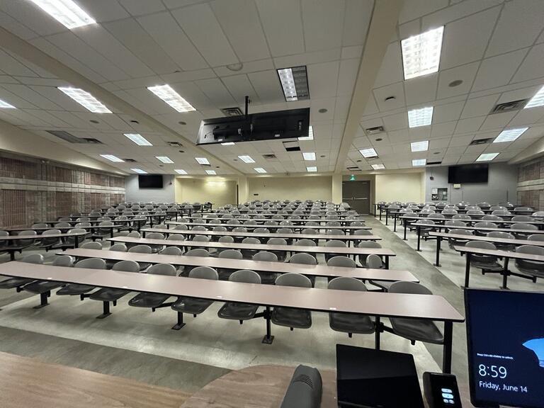 View of the classroom from the instructor's podium. There are 10 rows of long tables with swivel chairs for students, facing the front of the room. The entryway is located at the back on either side. This classroom is accessible for instructors.