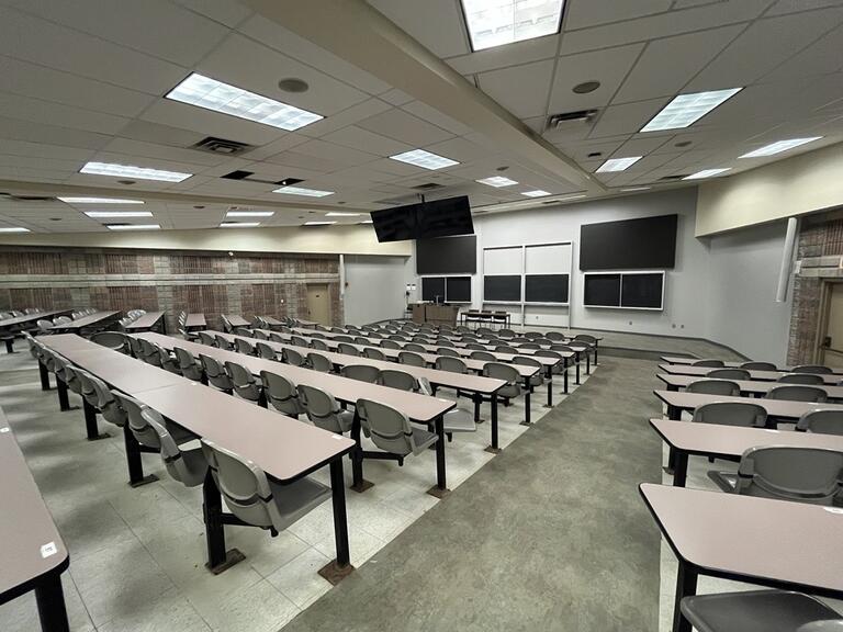 View of the classroom from a student seat near the rear of the room. In front are multiple empty chairs. The instructor podium is at the front left. There are 6 blackboards, and 2 video wall displays at the front of the room.