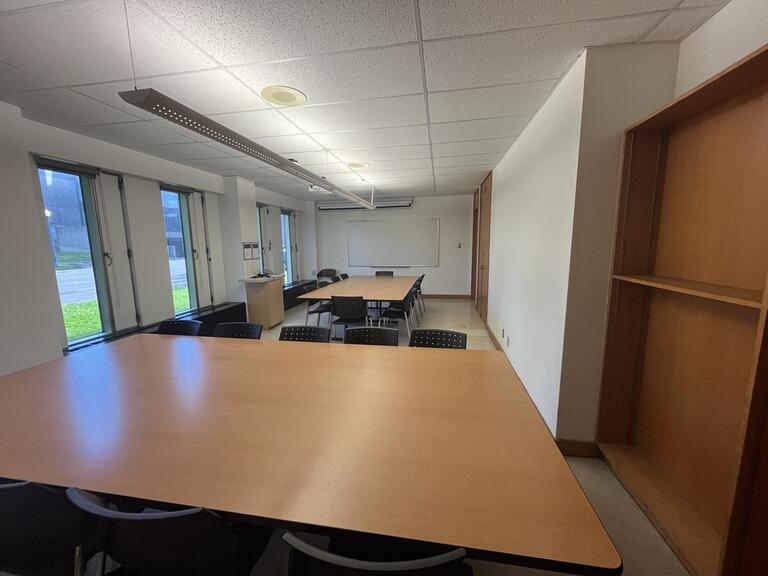 View of the classroom from a student seat near the rear of the room. In front are multiple empty chairs around a large table. The instructor’s podium is located at the front of the room on the left. A digital projector on the ceiling, projects onto a screen that extends from the ceiling. This classroom is accessible for students.