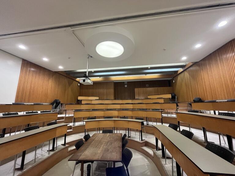 View of the classroom from the instructor’s podium. There are multiple rows of long curved tables with chairs for students. This classroom is not accessible for instructors.