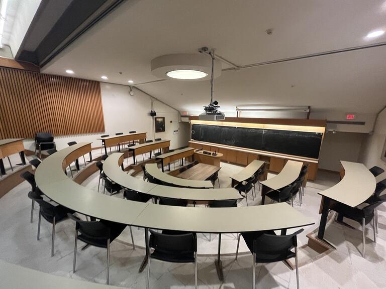 View of the classroom from a student seat near the rear of the room. In front are multiple rows of empty tables and chairs. The instructor’s podium is located at the front of the room on the left. A long blackboard is located on the front wall of the classroom. A digital projector on the ceiling, projects onto a screen that extends from the ceiling. This classroom is not accessible for students.