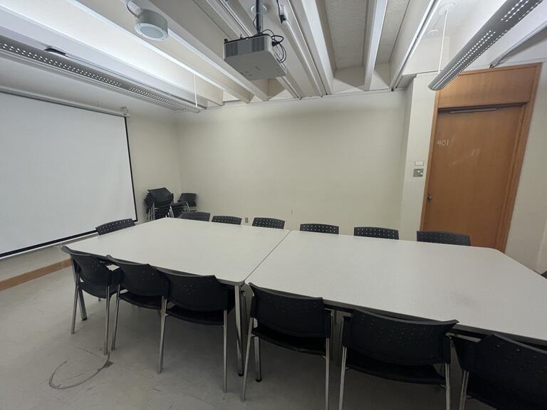 View of the classroom from the instructor’s podium. There are multiple chairs for students, arranged around a large table in the centre of the classroom. A projector on the ceiling projects onto a screen that extends from the ceiling. This classroom is accessible for instructors.