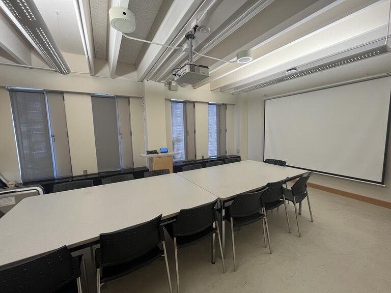 View of the classroom from a student seat near the rear of the room. In front are multiple empty chairs around a large table. The instructor’s podium is located at the front of the room on the right. A blackboard is located on the front wall of the room. This classroom is accessible for students.