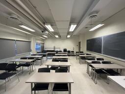View of the classroom from the instructor’s podium. There are rows of movable tables with chairs for students. Blackboards are located on the right wall of the room. The entryway door is located to the right at the back of the classroom. This classroom is accessible for instructors.