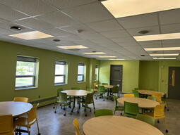View of the classroom from the instructor’s podium. There are multiple round tables with chairs for students. Two projectors on the ceiling project onto screens on the right hand side of the room. This classroom is accessible for instructors.