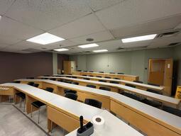 View of the classroom from the instructor’s podium. There are rows of long tables with chairs for students. The row at the back of the classroom is on a raised platform. The entryway door is located at the back of the room, on the left side. This room is 