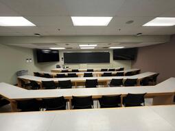 View of the classroom from a student seat near the rear of the room. In front are empty rows of tables and chairs. The instructor podium is at the front of the room on the left. The front of the classroom has a large blackboard in the middle of the front of the room, with two TV's on either side.