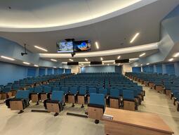 Instructor’s view of the classroom while standing at the front of the room. Seats are arranged in rows and are stationary. There are screens on the ceiling which displays media, mirroring the video wall at the front of the room. There are multiple exits in this room.