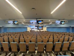This image depicts the students view when sitting at the rear of the room. There are rows of seating with tablet arms. The room slopes downwards towards the front of the room. There is accessible seating at the front and rear of the room. The instructor’s podium is in the front left.