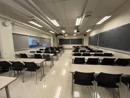 View of the classroom from a student seat near the rear of the room. In front are empty rows of tables and chairs. The instructor podium is at the front of the room on the left. The front of the classroom has two blackboards across the front wall, blackboards are also located on right side of the classroom. A digital projector on the ceiling, projects onto a screen that extends from the ceiling. This classroom is accessible for students.