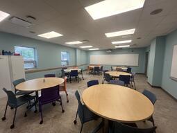 View of the classroom from the instructor’s podium. There are multiple round tables with chairs for students. A projector projects onto a screen, located at the back of the room. This classroom is accessible for instructors.