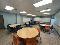 View of the classroom from a student seat near the rear of the room. In front are multiple round tables with chairs. The instructor podium is at the front of the room on the left. Two projectors project onto screens that are located across the front wall, as well as the left wall (from the perspective of a student). This classroom is accessible for students.