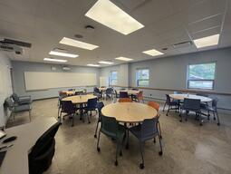 View of the classroom from the instructor’s podium. There are multiple round tables with chairs for students. A projector projects onto a screen, located at the back of the room. This classroom is accessible for instructors.