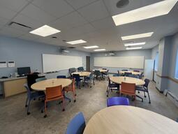 View of the classroom from a student seat near the rear of the room. In front are multiple round tables and blue and purple chairs. The instructor podium is at the front of the room on the left. The wall in front and to the left are covered in long whiteboards. On each wall a digital projector projects onto the whiteboard surface. This classroom is accessible for both instructors and students.