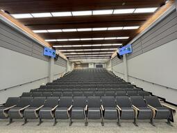 View of the classroom from the instructor’s podium. There are multiple rows of chairs for students. Chairs are arranged in rows with tiered steps to access the middle and back area of the room.