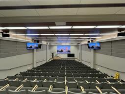 View of the classroom from a student seat near the rear of the room. In front are multiple empty rows of chairs. The instructor podium is at the front of the room on the left. The front of the classroom has a moveable blackboard and a video wall. This classroom is accessible for students.