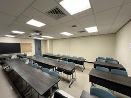 View of the classroom from the instructors podium. There are three rows of tables and chairs, with exits at the left of the room. 