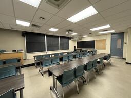 View of the classroom from a student seat near the rear of the room. In front are three empty rows of tables and chairs. The instructor podium is at the front of the room on the left. The front of the classroom has a three large blackboard panels in the middle of the front wall. A digital projector on the ceiling, projects onto a screen that extends from the ceiling. This classroom is accessible for students.