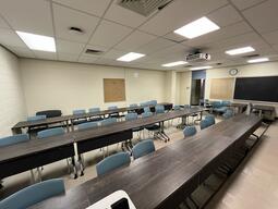 This image shows the instructors view in the room. The are three rows of desks and chairs that are both portable. At the back of the room there is a cork board on the wall. The exit is on the right side of the room.
