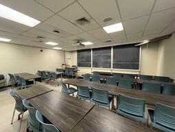 View of the classroom from a student seat near the rear of the room. In front are four empty rows of tables and chairs. The instructor podium is at the front of the room on the left. The front of the classroom has a three large blackboard panels in the middle of the front wall. A digital projector on the ceiling, projects onto a screen that extends from the ceiling. This classroom is accessible for students.