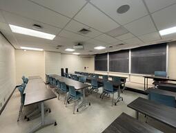 View of the classroom from a student seat near the rear of the room. In front are four empty rows of tables and chairs. The instructor podium is at the front of the room on the right. The front of the classroom has a three large blackboard panels in the middle of the front wall. A digital projector on the ceiling, projects onto a screen that extends from the ceiling. This classroom is accessible for students.