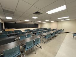 View of the classroom from a student seat near the rear of the room. In front are four empty rows of tables and chairs. The instructor podium is at the front of the room on the left. The front of the classroom has a three large blackboard panels in the middle of the front wall. A digital projector on the ceiling, projects onto a screen that extends from the ceiling. This classroom is accessible for students.