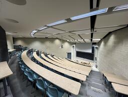 View of the classroom from a student seat near the rear of the room. In front are multiple empty rows of tables and chairs. The instructor podium is at the front of the room on the left. The front of the classroom has a large sliding blackboard panel on the front wall. This classroom is not accessible for students.