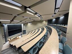 View of the classroom from a student seat near the rear of the room. In front are multiple empty rows of tables and chairs. The instructor podium is at the front of the room on the left. The front of the classroom has a large sliding blackboard panel on the front wall. This classroom is not accessible for students.