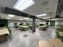 View from the instructors podium at the front of the room. There are tables with 8 chairs each throughout the room. The room is accessible for students and the instructor. Whiteboards cover all walls in the room. There are projectors throughout the room at multiple points which project onto the whiteboards.