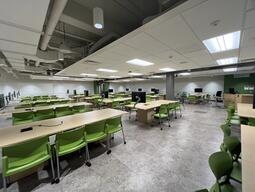 View of the classroom from a student seat. The podium is in the corner of the room and has whiteboards behind it. There is a computer at the podium. The podium is height adjustable and is accessible for instructors.