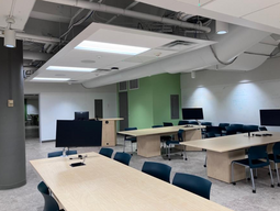 Student’s view while sitting at a table in the center of the room. The instructor’s podium is visible on the left. Tables seat 8 students each and the chairs are moveable. Each table has a monitor. The classroom is accessible for students.