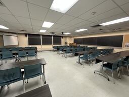 View of the classroom from a student seat near the rear of the room. In front are four empty rows of tables and chairs. The instructor podium is at the front of the room on the right. Three blackboard panels are located on the front wall. A projector on the ceiling projects onto a screen that extends from the ceiling. This classroom is accessible for students.