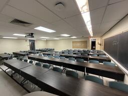 View of the classroom from the instructor’s podium. There are eight rows of long tables with chairs for students. The entryway door is located on the left side of the classroom. This classroom is not accessible for instructors.