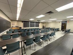 View of the classroom from a student seat near the rear of the room. In front are multiple empty rows of tables and chairs. The instructor podium is at the front of the room on the left. Three large sliding blackboard panels are located on the front wall. A projector on the ceiling projects onto a screen that extends from the ceiling. This classroom is accessible for students.