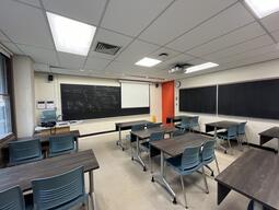 View of the classroom from a student seat near the rear of the room. In front are multiple empty rows of tables and chairs. The instructor podium is at the front of the room on the left. Two blackboard panels are located on the front wall. A projector on the ceiling projects onto a screen that extends from the ceiling. This classroom is accessible for students.