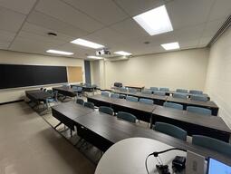 View of the classroom from the instructor’s podium. There are multiple tables with chairs for students, arranged in rows facing the front of the room. The entryway door is located on the left side of the classroom. This classroom is not accessible for instructors.