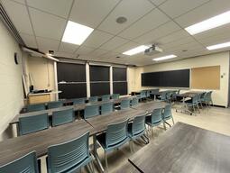 View of the classroom from a student seat near the rear of the room. In front are multiple empty rows of tables and chairs. The instructor podium is at the front of the room on the left. Three large sliding blackboard panels are located on the front wall. A projector on the ceiling projects onto a screen that extends from the ceiling. This classroom is accessible for students.