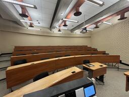 View of the classroom from the instructor’s podium. There are rows of long curved tables and chairs for students. This classroom has tiered steps, for access to the middle and back areas of the classroom. This classroom is accessible for instructors.