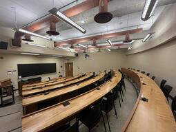 View of the classroom from a student seat near the rear of the room. In front are multiple empty rows of tables and chairs. The instructor’s podium is at the front of the room on the left. A long blackboard is located on the front wall. A projector on the ceiling projects onto a screen that extends from the ceiling. The entryway doors are located at the front of the room on the left and right. The front area of this classroom is accessible for students.