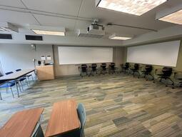 View of the classroom from a student seat near the rear of the room. In front are multiple empty chairs with tables attached. The instructor’s podium is located at the front of the room on the right. A projector on the ceiling projects onto a screen that extends from the ceiling, at the front of the room. This classroom is accessible for students.