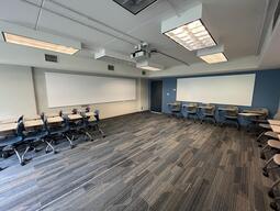 View of the classroom from the instructor’s podium. There are multiple tables with chairs attached for students, which can be arranged according to instructor’s preference. A whiteboard is located at the back of the room. The entryway door is located at the front of the room on the right. This classroom is accessible for instructors.